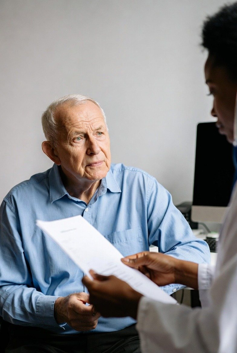 An older patient looks up at his doctor, who is reading a printed page of his records